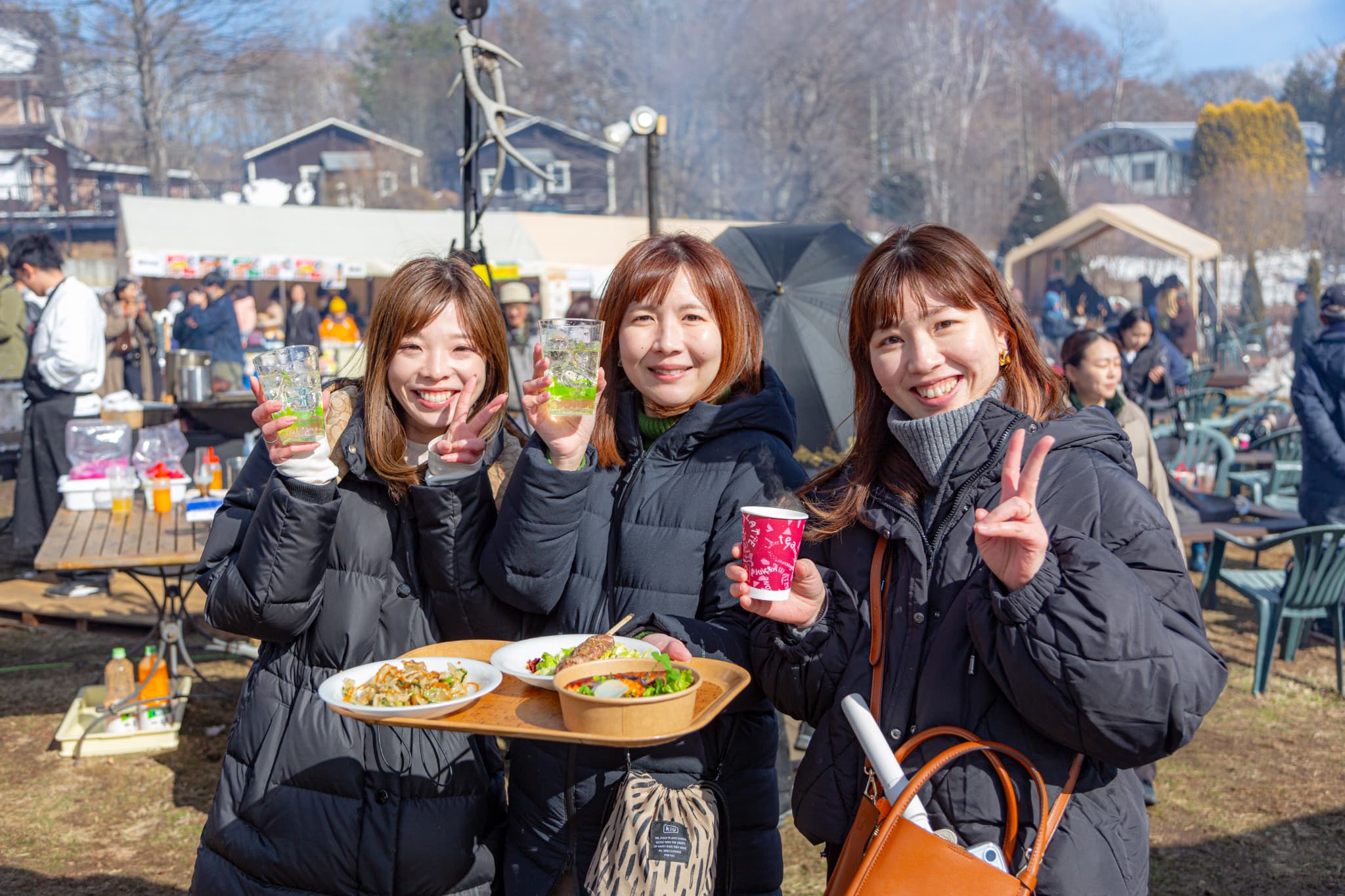 屋外で紙コップや料理を手に持ちカメラ目線でピースする女性3名